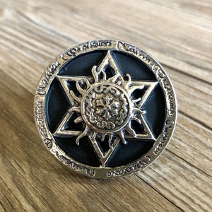 May include: A silver-colored, circular pendant with a black center. The pendant features a six-pointed star design with a sun-like emblem in the center. The outer edge has an engraved pattern. The pendant is likely a piece of jewelry or a decorative item.