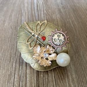 May include: A gold-toned metal brooch with a dragonfly, a flower, a gear, and a pearl. The dragonfly has red accents and the flower has a clear rhinestone in the center.