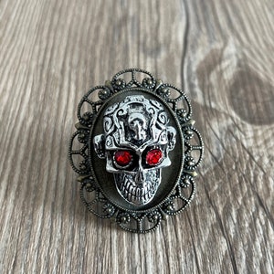 May include: A silver skull ring with red gem eyes set in an ornate, oval-shaped frame. The frame is made of a dark metal with a filigree design.