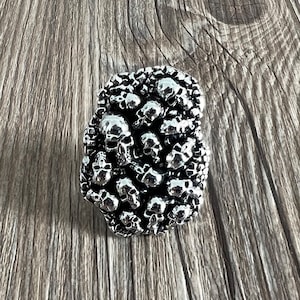 Könnte beinhalten: Eine silberfarbene, rechteckige Gürtelschnalle, die dicht mit kleinen Schädelformen bedeckt ist. Die Schädel sind mit Augenhöhlen, Nasen und Zähnen detailliert. Die Schnalle ist vor einem Holzgrund platziert.