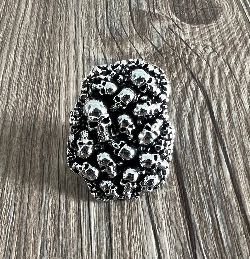 K&ouml;nnte beinhalten: Eine silberfarbene, rechteckige G&uuml;rtelschnalle, die dicht mit kleinen Sch&auml;delformen bedeckt ist. Die Sch&auml;del sind mit Augenh&ouml;hlen, Nasen und Z&auml;hnen detailliert. Die Schnalle ist vor einem Holzgrund platziert.