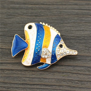May include: A gold-tone brooch in the shape of a blue, white, and orange fish with rhinestones. The fish has a blue tail and a white body with blue and orange stripes.