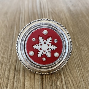 Puede incluir: Un pomo de copo de nieve plateado y rojo con acentos de perlas blancas. El pomo es redondo y tiene un diseño de copo de nieve en relieve.