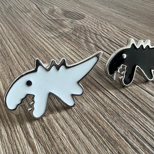 Może przedstawiać: Dwa metalowe przypinki w kształcie dinozaura, jedna czarna, a druga biała, z srebrnym wykończeniem. Przypinki mają kształt dinozaura z zębatym uśmiechem.