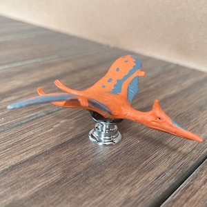May include: A cabinet knob with a Pterodactyl design. The knob has an orange body with grey wing accents and a chrome base. The dinosaur is in a flying pose, adding a unique touch to furniture.