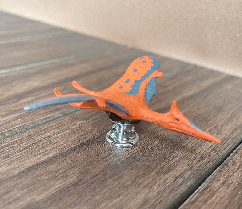 May include: A cabinet knob with a Pterodactyl design. The knob has an orange body with grey wing accents and a chrome base. The dinosaur is in a flying pose, adding a unique touch to furniture.