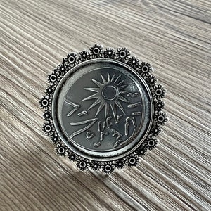 以下が含まれることがあります： 太陽とスタイリッシュな波が描かれた円形のデザインのシルバーリング。リングは、小さなシルバーの花の装飾的な縁で囲まれています。