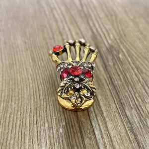 K&ouml;nnte beinhalten: Goldfarbene Skelett-Hand-Brosche mit roten und klaren Strasssteinen.