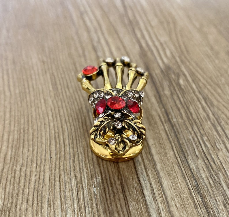 K&ouml;nnte beinhalten: Goldfarbene Skelett-Hand-Brosche mit roten und klaren Strasssteinen.