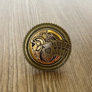 Puede incluir: Un anillo de oro y bronce con un diseño steampunk que presenta un mecanismo de engranaje y cartas de juego con el as de corazones, rey, reina, jota y diez de corazones.