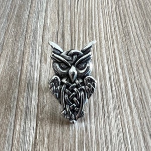 Op de afbeelding: Een zilveren uilhanger met een Keltisch knoopontwerp. De ogen van de uil zijn gedetailleerd en de vleugels zijn uitgespreid.