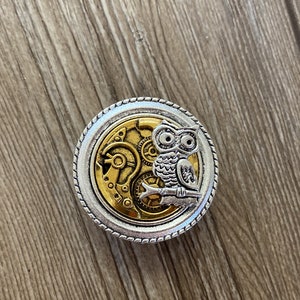 May include: A silver and gold round metal pin with a detailed image of a steampunk owl with gears and cogs.