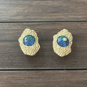 Könnte beinhalten: Ein Paar goldfarbene Ohrringe mit einer strukturierten Oberfläche. Jeder Ohrring zeigt ein blaues und grünes Emaille-Design mit einer weißen Perle und einem Halbmond.