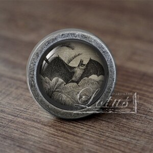 May include: A silver-toned cabinet knob with a glass insert featuring a black and white illustration of a bat flying over a field of grass.