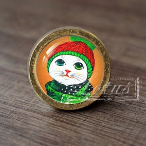 May include: A round, brass cabinet knob with a glass insert featuring a white cat wearing a red and green knitted hat and a green scarf with white polka dots.