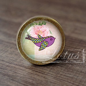 Può includere: Un pomello rotondo per armadio color oro con un inserto in vetro che presenta un uccello viola e verde su uno sfondo di fiori rosa.