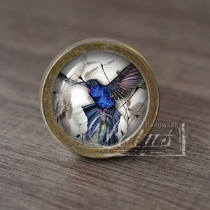 Peut inclure: Bouton de meuble rond avec une image détaillée d'un colibri bleu aux ailes déployées. Le bouton a un bord en métal couleur laiton et un centre transparent en forme de dôme. Le colibri est placé sur un fond d'éléments floraux blancs et gris.