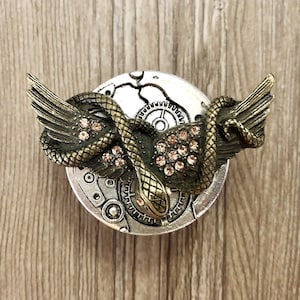 Könnte beinhalten: Ein silberner und bronzenfarbener Anhänger im Steampunk-Stil mit einer Schlange mit Flügeln, die um ein Uhrwerk mit Zahnrädern und Strasssteinen gewickelt ist.