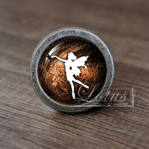 Peut inclure: Bouton de meuble décoratif avec une base en métal argenté. Le bouton présente un centre rond de couleur bronze avec une silhouette blanche de fée. Le mot "Lotus" est visible sur le bouton.
