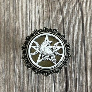 May include: A silver crow sits atop a pentagram with three crescent moons, all within a circular frame of bronze with a floral design.