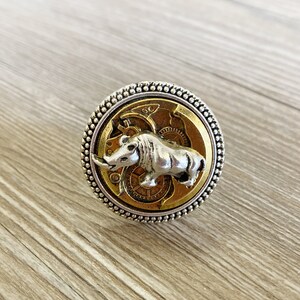May include: A silver ring with a gold-toned center featuring a detailed gear design and a silver rhinoceros figurine.