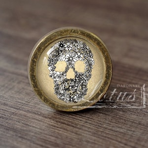 Puede incluir: Pomo de armario redondo con un diseño de calavera hecho de flores blancas y negras. El pomo tiene una base de metal color latón y una parte superior de vidrio o resina transparente. Se ven las cuencas de los ojos y la nariz de la calavera. El pomo está sobre una superficie de madera.
