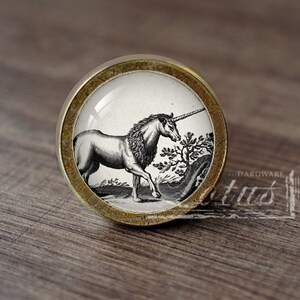 May include: A round, brass-colored cabinet knob with a glass insert featuring a black and white illustration of a unicorn. The unicorn is standing in a grassy field with a tree behind it.