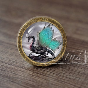 Op de afbeelding: Een ronde, messingkleurige knop met een glazen inzetstuk met een zwarte draak met turquoise vleugels. De draak is geplaatst tegen een achtergrond van een bewolkte hemel.