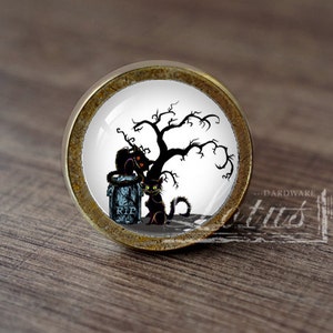 Pode incluir: Um botão de armário redondo de cor bronze com um inserto de cerâmica branca que apresenta uma silhueta de gato preto sob uma árvore nua com uma lápide que diz "RIP".