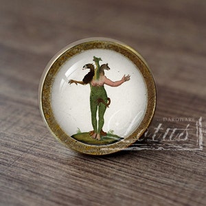 May include: A round cabinet knob with a brass-colored rim and a clear glass center. The center features a vintage illustration of a mythical creature with three heads, green scales, and outstretched arms. The knob is set against a wooden surface.