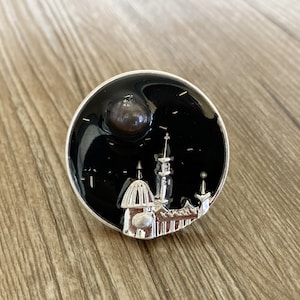 Puede incluir: Broche redondo plateado con fondo de esmalte negro que representa un paisaje urbano. Una perla oscura está colocada en la parte superior. El broche presenta un diseño arquitectónico detallado.