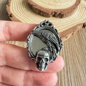 May include: A silver-toned pendant featuring a raven perched above a skull within an oval frame. The raven's detailed feathers and the skull's intricate design are prominent. The pendant is held in a hand, showcasing its size and detail.