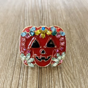 以下が含まれることがあります： 赤、金、そしてマルチカラーの宝石で飾られたカボチャのブローチ。カボチャは、大きく笑った黒い顔と2つの目を持っています。ブローチはラインストーンやその他の宝石で飾られています。