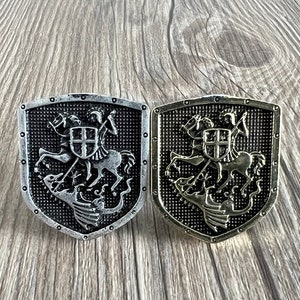 May include: Two silver-toned metal belt buckles with a shield design. Each shield features a knight riding a horse and slaying a dragon. The shields have a black background and a textured surface.