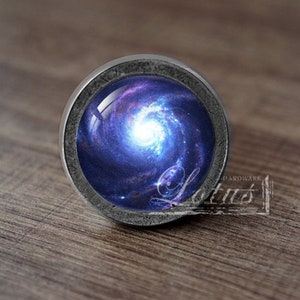 Puede incluir: Un pomo plateado con un diseño de galaxia. La galaxia es un remolino azul y morado con un centro blanco.