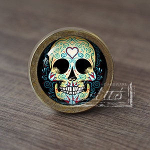 May include: A round, brass-colored cabinet knob with a colorful, detailed sugar skull design. The skull is yellow, blue, red, and white with black eyes and a small white heart on the forehead.
