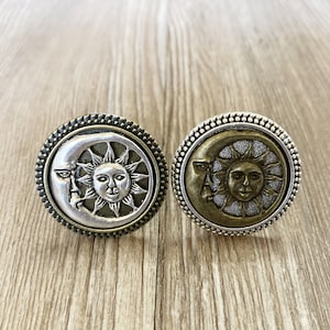 May include: Two decorative metal rings with a sun and moon design. The rings are silver and gold in color and have a textured finish.