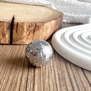 May include: A hammered silver sphere with a textured surface. The sphere is displayed on a wooden surface, next to a white, circular, tiered object and a wooden slice. The sphere reflects light, creating a metallic sheen.