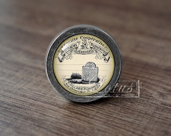 May include: A silver knob with a black and white image of a vintage pharmacy label. The label reads "Societe Cooperative Pharmacies Populaires Liegeoises H. Gillard Pharmacien Rue d'Amercoeur 71, Liege".