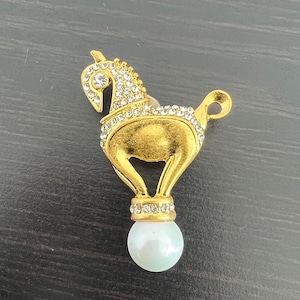 Può includere: Spilla a forma di cavallo color oro con dettagli in strass trasparenti. Il cavallo è in piedi su un globo perlato verde chiaro. La spilla ha un'estetica vintage.