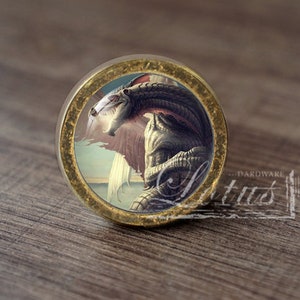 Op de afbeelding: Een ronde, gouden kastknop met een gedetailleerd, sierlijk ontwerp. De knop heeft een kleurrijk beeld van een draak met uitgespreide vleugels.