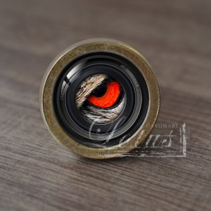 Puede incluir: Un pomo de armario redondo de color latón con una lente de cámara negra en el centro. La lente tiene un ojo rojo en el centro, rodeado de una imagen en blanco y negro de piel.