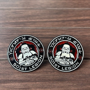Könnte beinhalten: Zwei schwarze und rote Emaille-Pins mit einem weißen Stormtrooper-Design. Die Pins zeigen den Text "501st Legion" und Aurebesh-Text am Rand.