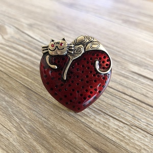May include: A heart-shaped brooch with a red, perforated surface and a gold-colored cat design. The cat has red gem eyes and is curled up on the heart. The brooch is made of metal and has a vintage aesthetic.