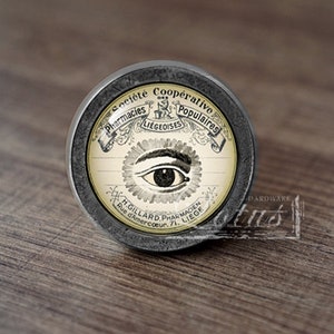 Pode incluir: Um botão de armário redondo, de cor prateada, com um design de estilo vintage. O botão apresenta uma imagem em preto e branco de um olho com o texto "Societe Cooperative Pharmacies Populaires Liegeoises H. Gillard Pharmacien Rue d'Amercoeur, 1, Liege" em torno do olho.