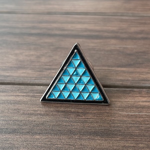 Może przedstawiać: Srebrna, trójkątna spinka z niebieskim i czarnym geometrycznym wzorem.