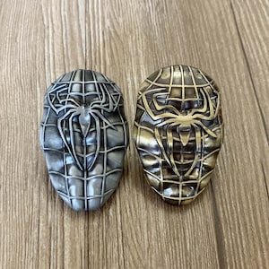 May include: Two metal keychains featuring a stylized spider web design with a spider in the center. One keychain is silver and the other is gold.