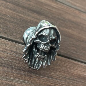 Op de afbeelding: Zilverkleurige kastknop in de vorm van een schedel met een kap. De schedel heeft gedetailleerde gelaatstrekken en is gemonteerd op een ronde basis. De metalen knop heeft een gotische of macabere uitstraling voor meubels of decoratie.