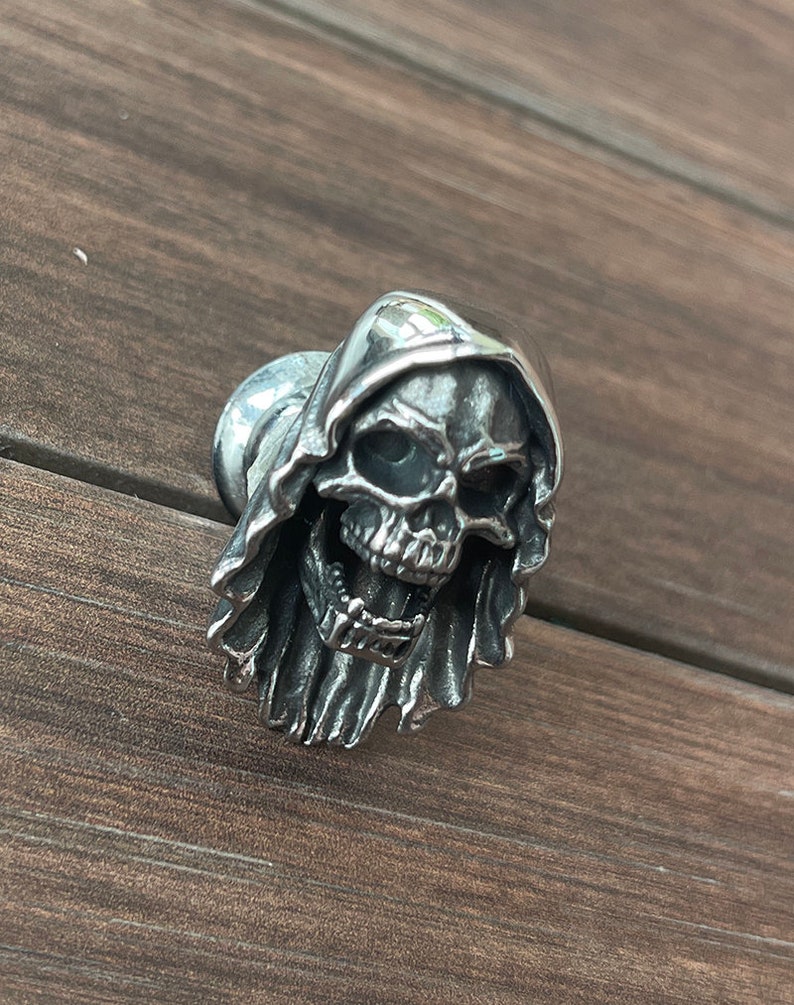 Op de afbeelding: Zilverkleurige kastknop in de vorm van een schedel met een kap. De schedel heeft gedetailleerde gelaatstrekken en is gemonteerd op een ronde basis. De metalen knop heeft een gotische of macabere uitstraling voor meubels of decoratie.