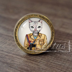 May include: A round, brass-colored cabinet knob with a white cat wearing a brown jacket and a red scarf. The cat has a mustache and a serious expression.